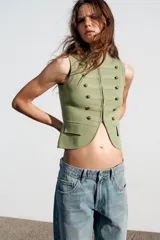 Chaleco de punto en color verde claro, con cuello redondo y diseño sin mangas. Presenta un estilo militar con detalles de alamares, botones decorativos dorados en el frente y bolsillos falsos con tapeta. Cierre frontal con corchetes ocultos.