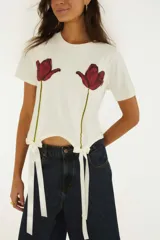 Remera blanca de manga corta y cuello redondo, confeccionada en algodón, con estampado frontal de dos tulipanes rojos y detalles de lazos de satén en el dobladillo.
