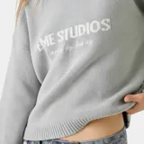 Suéter de punto grueso color gris, con corte holgado y ligeramente corto. Presenta cuello redondo, puños y ruedo acanalados, con el logo de la marca y la frase 'curated by ben ice' tejidos en jacquard en el frente.