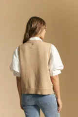 Chaleco tejido sin mangas color blanco roto, con cuello redondo y cartera frontal de botones. Presenta un punto calado con textura.