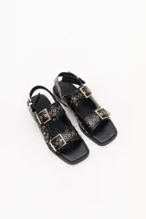 Sandalias negras de cuero con suela plana, dos tiras anchas con hebillas doradas y apliques metálicos.