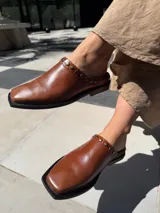 Zuecos de cuero marrón con diseño destalonado, punta cuadrada y detalles de tachas metálicas en el empeine.