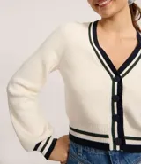 Cardigan corto de punto color crema con escote en V ribeteado en negro y blanco. Presenta cierre frontal de botones negros y puños con el mismo ribete a rayas. Las mangas son largas y ligeramente abullonadas.