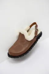 Zueco de cuero color suela con interior forrado en piel sintética blanca. Presenta una tira trasera ajustable con hebilla metálica y suela de goma negra con relieve.