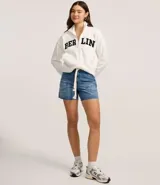 Chaqueta deportiva blanca con cierre frontal, cuello alto y puños ajustados. Presenta la inscripción "BERLIN" en letras negras y gruesas en el pecho.