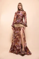 Vestido largo de mujer estilo boho en tono marrón, con estampado paisley y floral, cuello alto, abertura frontal en el escote y mangas largas abullonadas. Cuenta con detalles de aplicaciones de pedrería y flecos en la cintura, y falda larga con tajo lateral.