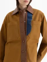 Chaqueta color marrón confeccionada en gabardina, con cuello y cartera de cuero marrón. Presenta broches a tono de cuero en la cartera y puños, bolsillos plaqué y sastreros en el delantero. Incluye volados de denim azul que aportan un toque original al diseño.