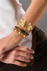 Conjunto de dos pulseras y un brazalete dorado. El brazalete rígido presenta un diseño de flores troqueladas con textura, mientras que las pulseras elásticas combinan cuentas metálicas doradas y cilindros de resina en tonos marrones.