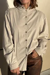 Camisa de pana color beige, con cuello alto con volados, cierre frontal de botones y mangas largas con puños abotonados.