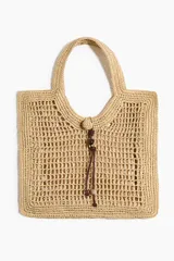 Cartera tote de paja trenzada con diseño calado, asas de hombro y cierre con cordón y cuentas decorativas.