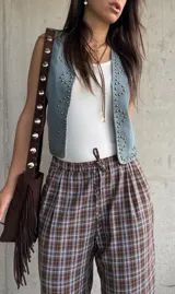 Chaleco de jean color celeste claro, con escote en V, sin mangas y decorado con tachas metálicas doradas distribuidas en los bordes y formando diseños en los paneles frontales.
