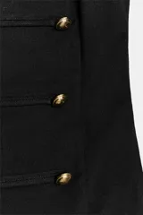 Vestido corto de estilo militar en color negro, con cuello redondo, hombreras y manga sisa. Presenta un diseño frontal con falsos botones dorados dispuestos en filas horizontales. Cierre lateral con cremallera oculta y abertura en la espalda con botón.