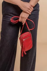 Bandolera porta celular color negro, confeccionada en símil cuero con textura granulada. Presenta un compartimento principal con cierre, un bolsillo frontal con cierre y detalle de borla decorativa, y correa regulable y desmontable.