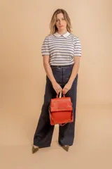 Mochila tipo cartera con tapa, confeccionada en símil cuero color suela. Cuenta con doble compartimento, división interna, cierre de seguridad y bolsillo trasero con cierre. Incluye una correa frontal decorativa con tachas metálicas.