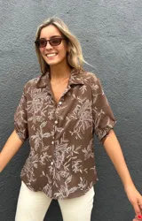 Camisa de corte recto y manga corta con cuello clásico y cierre frontal de botones. Presenta un estampado floral en color blanco sobre una base de color chocolate.