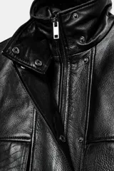 Chaqueta de piel negra con cuello alto, manga larga y cierre frontal con cremallera y botones a presión ocultos bajo solapa. Presenta bolsillos de plastrón con solapa en el frente y forro interior a tono.