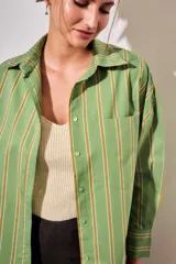 Camisa de corte oversize con estampado de rayas verticales en tonos verde, blanco y ocre. Presenta cuello clásico, cierre frontal con botones, bolsillo en el pecho y mangas largas con puños abotonados.