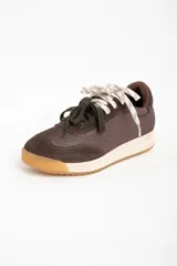 Zapatilla deportiva de perfil bajo, color marrón chocolate, con paneles de gamuza y malla texturizada. Presenta una entresuela de color crema y suela de goma color caramelo. Se ata con dos pares de cordones: uno blanco trenzado y otro verde oscuro.