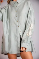 Camisa beige de corte amplio, con cuello clásico, cierre frontal con botones, mangas largas con puños abotonados y bordado floral en las mangas.