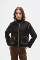 Chaqueta negra de poliéster con cuello a la base, cierre frontal con botones y dos bolsillos delanteros. Presenta detalles de bordado decorativo en color marrón en los bordes, cuello, puños y bolsillos.