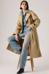 Trench coat largo de corte clásico en color beige, con diseño de doble botonadura, solapas amplias y cuello con detalle de textura. Presenta un estilo atemporal y estructurado.
