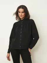 Camisa de denim negro con cuello alto tipo mao con volados, cierre frontal con botones metálicos y mangas largas con hombros ligeramente fruncidos.