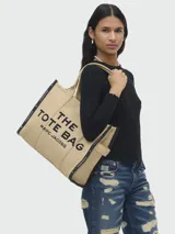 Bolso tipo tote de gran tamaño en color beige, confeccionado en lona resistente. Presenta el logo estampado en negro en el frente y detalles de cintas laterales con inscripciones. Cuenta con doble asa superior para llevar al hombro.