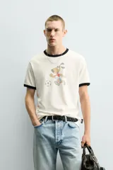 Camiseta de algodón color crudo con cuello y bordes de mangas en contraste negro. Presenta un estampado frontal de estilo vintage con el personaje Striker, mascota de la Copa Mundial de la FIFA 1994, jugando al fútbol.