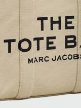 Bolso tipo tote bag de Marc Jacobs en color beige, confeccionado en lona resistente. Presenta el texto "THE TOTE BAG" y "MARC JACOBS" impreso en la parte frontal en color negro. Cuenta con asas superiores y una correa de hombro ajustable y extraíble, además de ribetes laterales negros con inscripciones.