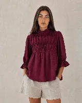 Blusa holgada color blanco, de manga por debajo del codo con elástico y volados en los puños. Presenta cuello alto con volados y diseño fruncido tipo nido de abeja hecho a mano en la parte superior delantera.