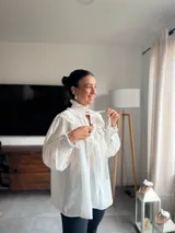 Blusa de color marrón con diseño romántico, cuello alto con lazo ajustable y volados tipo shabot en el frente. Presenta mangas largas abullonadas con puños ajustados y una textura suave y liviana.