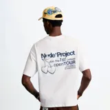 Remera oversize Nude Project de color blanco, de manga corta y cuello redondo. Presenta un estampado gráfico en la espalda con la inscripción "Nude Project Join The 1st Leisure openTOUR" y el dibujo de dos cerezas con forma de corazón.