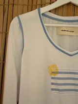 Remera de manga larga blanca con escote en V y vivos en color celeste. Presenta un bolsillo frontal con estampado de rayas celestes y un diseño circular bordado en color amarillo. Las mangas tienen un ligero fruncido en los hombros.