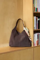 Cartera tipo tote de tamaño mediano, confeccionada en cuero suave color marrón oscuro. Presenta un diseño minimalista con costura central vertical y un pañuelo estampado anudado en una de las asas.