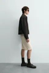 Bermuda de algodón color beige claro, de corte relajado y largo hasta la rodilla. Presenta cierre frontal con cremallera y botón, bolsillos delanteros y bolsillo cerillero, además de bolsillos traseros de plastrón.