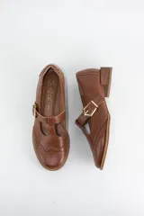 Zapato tipo guillermina estilo oxford, confeccionado en cuero negro con detalles de perforaciones decorativas en la puntera y cierre mediante tira con hebilla metálica dorada. Presenta un taco bajo cuadrado.