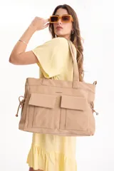 Bolso tipo tote color beige con dos bolsillos frontales grandes con solapa y cierre de broche, asas de hombro fijas y detalles de cordones ajustables en los laterales.