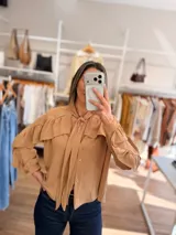 Camisa color tostado con volados en los hombros, mangas largas abullonadas y cuello con lazo para anudar. Presenta cierre frontal con botones y diseño de calce amplio.