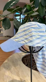 Gorro tipo gorra con visera rígida celeste y pañuelo integrado de tela a rayas blancas y celestes, con detalles de bordado a mano en color naranja.