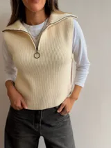 Chaleco de tejido de punto acanalado color manteca, con cuello alto, cierre frontal parcial con tirador circular y aberturas laterales con botones.