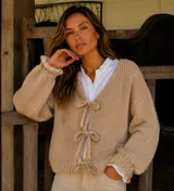 Cardigan de punto grueso en lana merino color beige, con escote en V y cierre frontal mediante lazos de tela. Presenta mangas largas voluminosas con puños acanalados.