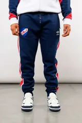 Pantalón deportivo tipo jogging de la marca Umbro, en color azul marino con bandas laterales en rojo y blanco. Cuenta con el escudo del Club Nacional de Football en el muslo derecho y el logo de Umbro en el izquierdo, además de cintura elástica ajustable con cordón y puños en los tobillos.