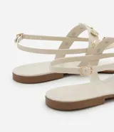 Sandalia plana color beige con tiras finas y tira central decorada con colgantes metálicos dorados en forma de conchas marinas. Cuenta con cierre de hebilla ajustable en el tobillo.