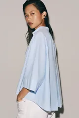 Camisa Zara Woman Collection de algodón con estampado de rayas verticales blancas y celestes. Presenta cuello perkins, mangas tres cuartos y un diseño asimétrico con cierre frontal cruzado mediante botones.