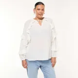 Blusa blanca de manga larga con textura de plumeti (pequeños puntos en relieve) y volantes decorativos verticales en el frente y en los puños. Presenta un escote en V con lazos para anudar en el cuello.