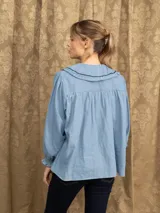 Camisa de jean azul con cuello babero, detalles de volados y bordados en contraste. Presenta mangas largas con puños elásticos y cierre frontal con botones.