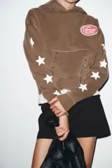 Sudadera marrón con capucha ajustable con cordones, manga larga con puños y bajo elástico. Presenta un estampado ovalado en el pecho con el texto "Texas USA" en rosa y azul, y un diseño de estrellas blancas en las mangas. El bolsillo canguro frontal tiene detalles de tachas y bordes deshilachados.