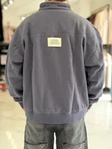 Buzo gris oscuro de corte oversize con cuello alto y cierre frontal parcial. Cuenta con mangas largas y puños elásticos, además de un pequeño bordado con texto en el pecho.