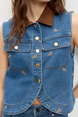 Chaleco de denim azul sin mangas, con cierre frontal de botones metálicos, bolsillos con solapa en el pecho y cuello camisero marrón de pana. Presenta bordados pequeños dispersos en color marrón.