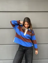 Buzo tipo rugby de algodón con diseño de rayas horizontales anchas en azul y marrón. Presenta cuello polo en color crema con cierre frontal de tres botones y mangas largas.